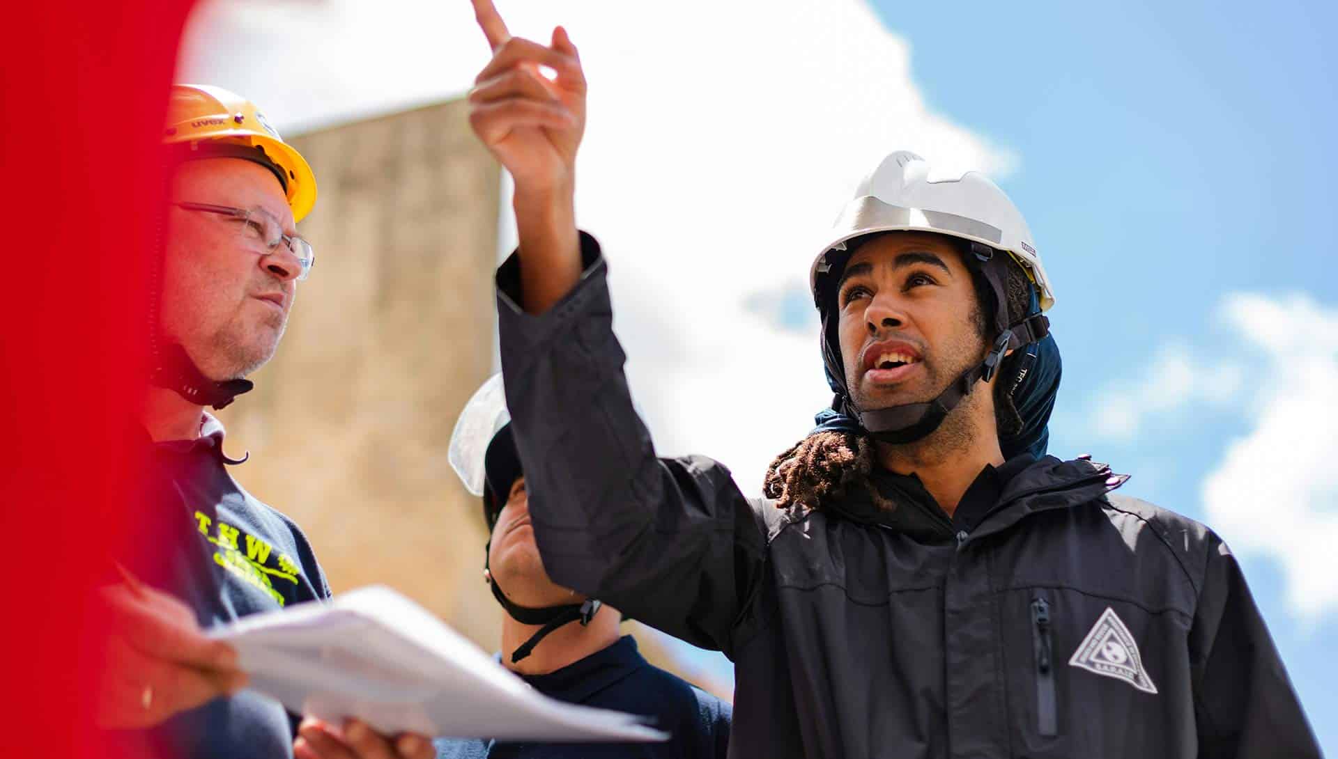 Ingeniero de construcción en casco y chaqueta negra discutiendo con supervisor en casco amarillo y ropa de trabajo en sitio de obra.